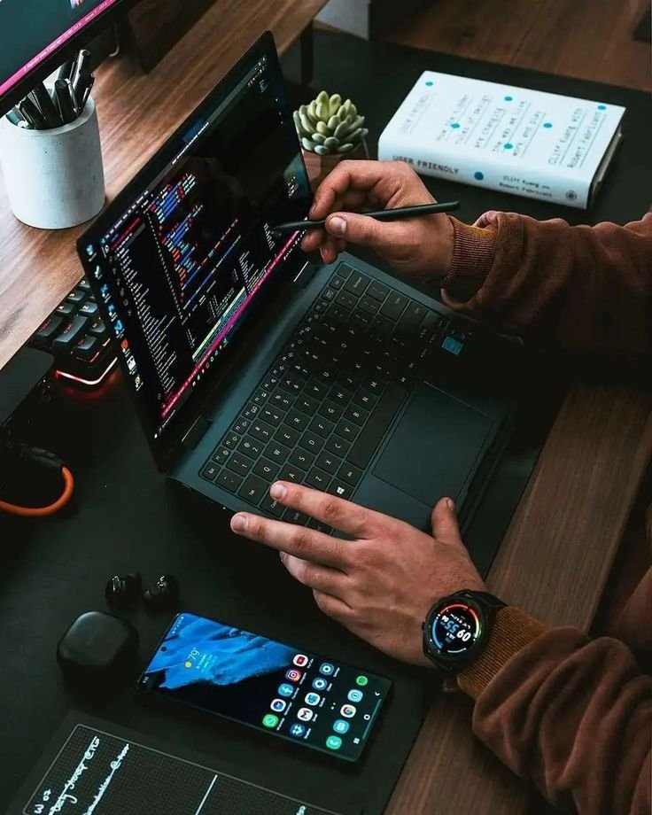 A person working on a laptop with a stylus, engaged in web development. The workspace includes a smartphone displaying multiple app icons, a smartwatch showing activity stats, wireless earbuds, and a book titled "User Friendly." The setting is a modern desk setup with various tech gadgets, highlighting a productive and tech-savvy environment.